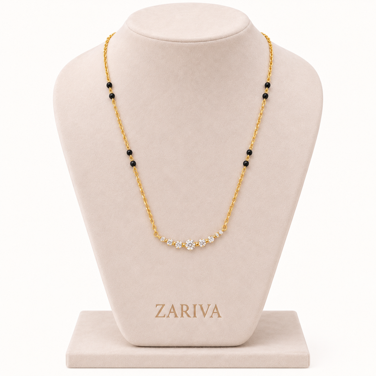 A gold chain necklace with a curved row of small clear stones at the center and small black bead accents spaced along the chain, displayed on a beige jewelry bust. The brand name ZARIVA is visible on the base of the bust.