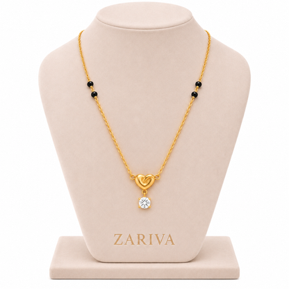 A gold necklace displayed on a beige jewelry bust. The necklace has a gold chain with small black beads and a pendant featuring a gold heart and a clear round gemstone.