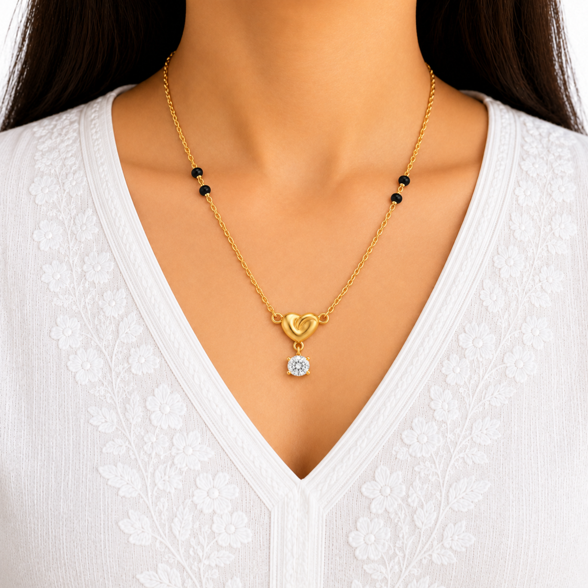 A close-up of a model wearing a delicate gold chain necklace with a heart-shaped pendant and small black beads, paired with a white lace V-neck top.