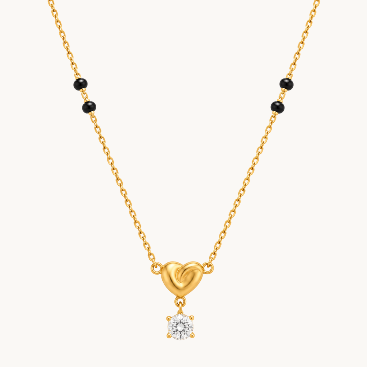 A gold chain necklace featuring a small heart-shaped pendant with a dangling clear gemstone, accented by small black beads on the chain, shown against a light off-white background.