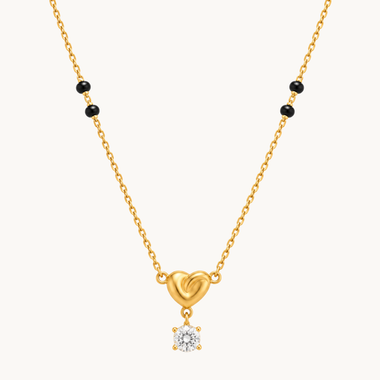 A gold chain necklace featuring a small heart-shaped pendant with a dangling clear gemstone, accented by small black beads on the chain, shown against a light off-white background.