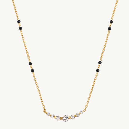 A delicate yellow gold chain necklace featuring small black round beads spaced along the chain and a curved central section set with clear gemstones, photographed on a solid off-white background.