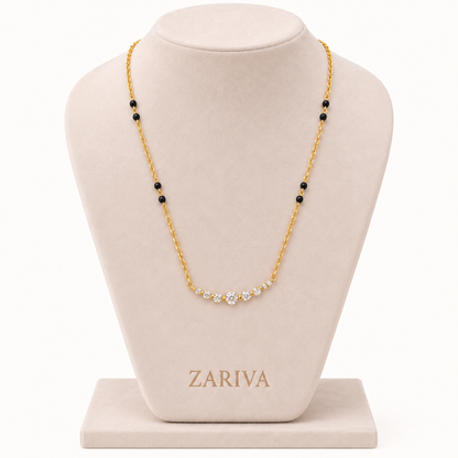 A gold chain necklace with a curved row of small clear stones at the center and small black bead accents spaced along the chain, displayed on a beige jewelry bust. The brand name ZARIVA is visible on the base of the bust.