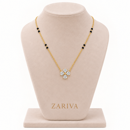 A gold necklace with a four-petal flower pendant set with clear stones, displayed on a beige jewelry bust. The necklace has small black bead accents along the chain.